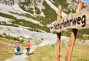 Familie wandert auf einem Naturlehrweg in der Schlick 2000, im Vordergrund steht ein hölzernes Wegschild vor alpiner Sommerlandschaft.