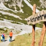 Familie wandert auf einem Naturlehrweg in der Schlick 2000, im Vordergrund steht ein hölzernes Wegschild vor alpiner Sommerlandschaft.