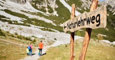 Familie wandert auf einem Naturlehrweg in der Schlick 2000, im Vordergrund steht ein hölzernes Wegschild vor alpiner Sommerlandschaft.