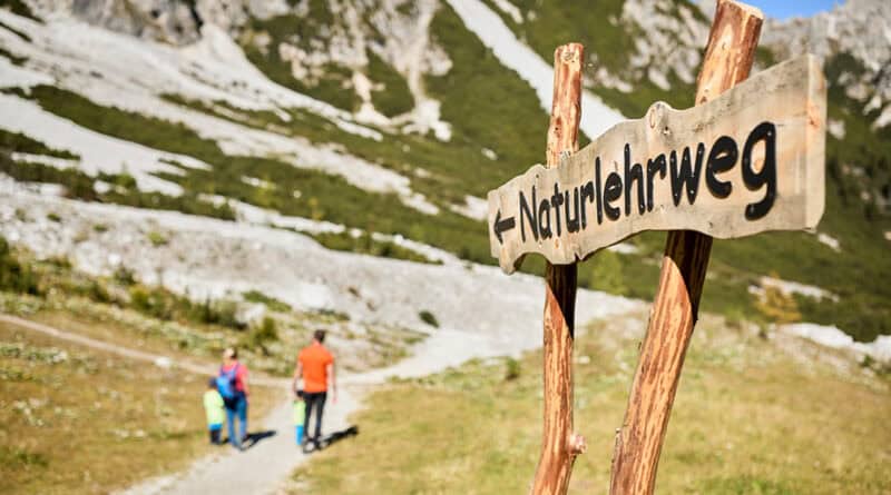 Familie wandert auf einem Naturlehrweg in der Schlick 2000, im Vordergrund steht ein hölzernes Wegschild vor alpiner Sommerlandschaft.