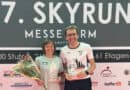 Annemarie Wilhelm und Dr. Christian Riedl stehen nach dem 17. SkyRun MesseTurm Frankfurt vor der Veranstaltungswand und präsentieren ihre Siegerpokale.