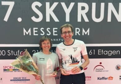 Annemarie Wilhelm und Dr. Christian Riedl stehen nach dem 17. SkyRun MesseTurm Frankfurt vor der Veranstaltungswand und präsentieren ihre Siegerpokale.
