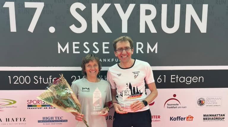 Annemarie Wilhelm und Dr. Christian Riedl stehen nach dem 17. SkyRun MesseTurm Frankfurt vor der Veranstaltungswand und präsentieren ihre Siegerpokale.