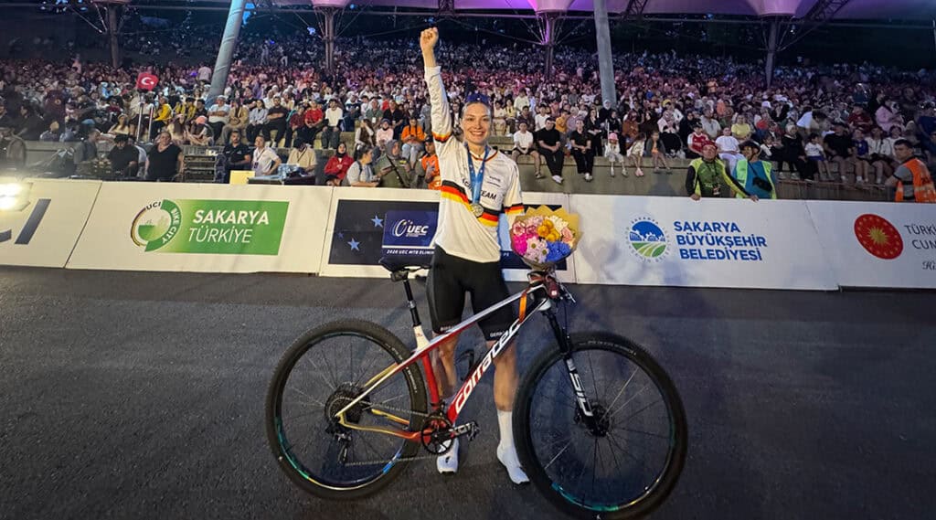 Marion Fromberger strahlt nach ihrem erfolgreichen Rennen in Sakarya und hebt die rechte Faust zum Sieg. In der linken Hand hält sie einen Blumenstrauß und ihr Corratec MTB.