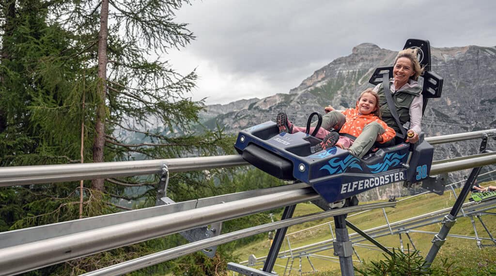 Eine Erwachsene und ein Kind fahren gemeinsam mit dem ELFERCOASTER auf einer Schienenrodelbahn vor der Bergkulisse des Stubaitals.