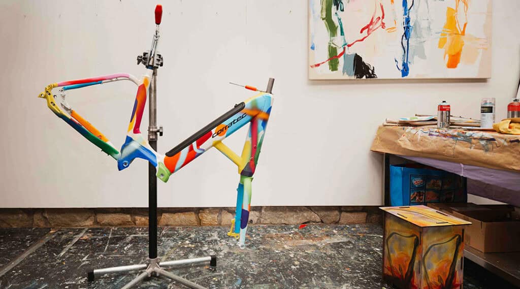 Bunt bemalter corratec LIFE S Fahrradrahmen auf einem Montageständer im Atelier.