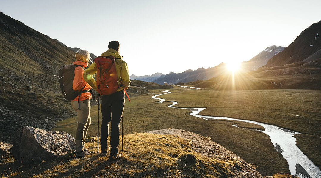 Herbst im Stubaital: Zwei Wanderer blicken im goldenen Abendlicht auf eine alpine Landschaft mit Bachlauf und Stubaier Bergen.