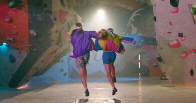 Zwei Musiker laufen gemeinsam durch eine Boulderhalle, umgeben von bunten Kletterwänden.