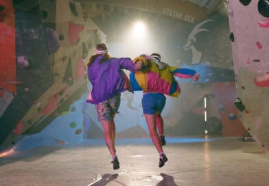Zwei Musiker laufen gemeinsam durch eine Boulderhalle, umgeben von bunten Kletterwänden.