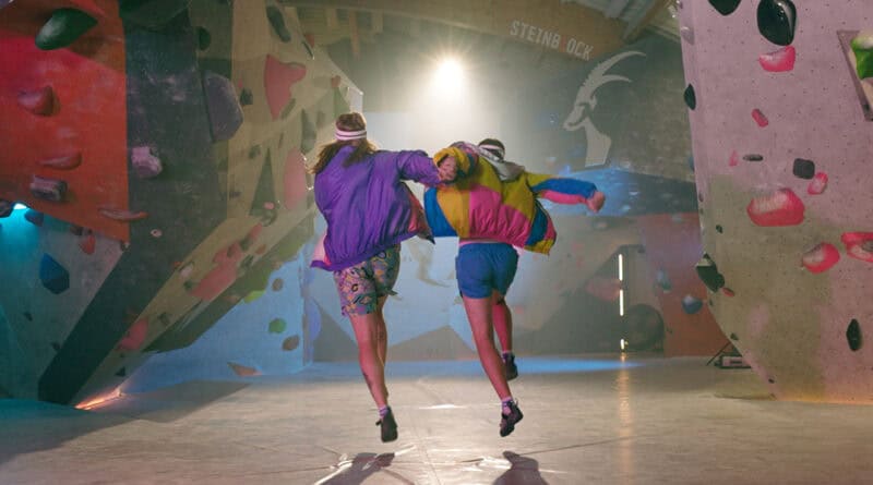 Zwei Musiker laufen gemeinsam durch eine Boulderhalle, umgeben von bunten Kletterwänden.