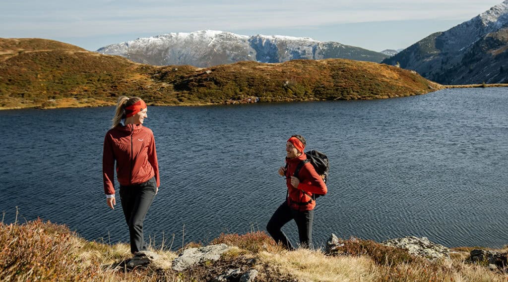 Zwei Wanderinnen in roter Outdoorbekleidung an einem Bergsee in der Region Hohe Salve, mit schneebedeckten Bergen im Hintergrund.