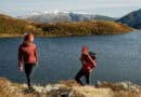 Zwei Wanderinnen in roter Outdoorbekleidung an einem Bergsee in der Region Hohe Salve, mit schneebedeckten Bergen im Hintergrund.