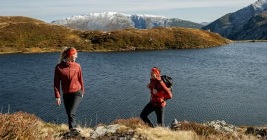 Zwei Wanderinnen in roter Outdoorbekleidung an einem Bergsee in der Region Hohe Salve, mit schneebedeckten Bergen im Hintergrund.