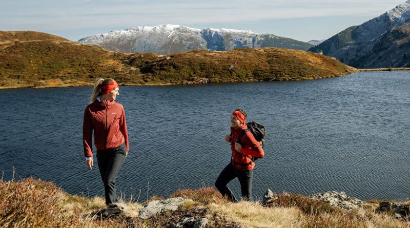 Zwei Wanderinnen in roter Outdoorbekleidung an einem Bergsee in der Region Hohe Salve, mit schneebedeckten Bergen im Hintergrund.