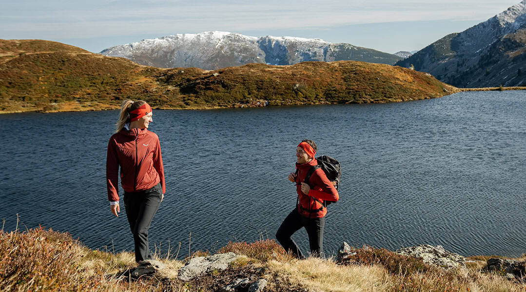 Zwei Wanderinnen in roter Outdoorbekleidung an einem Bergsee in der Region Hohe Salve, mit schneebedeckten Bergen im Hintergrund.