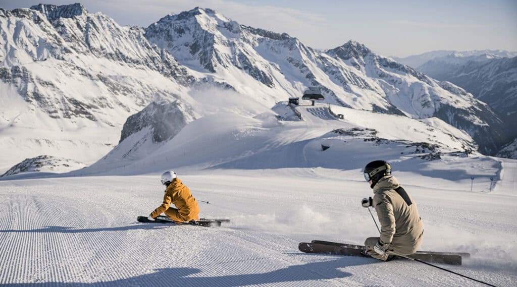 Zwei Skifahrer auf frisch präparierter Piste am Stubaier Gletscher vor verschneiter Bergkulisse.