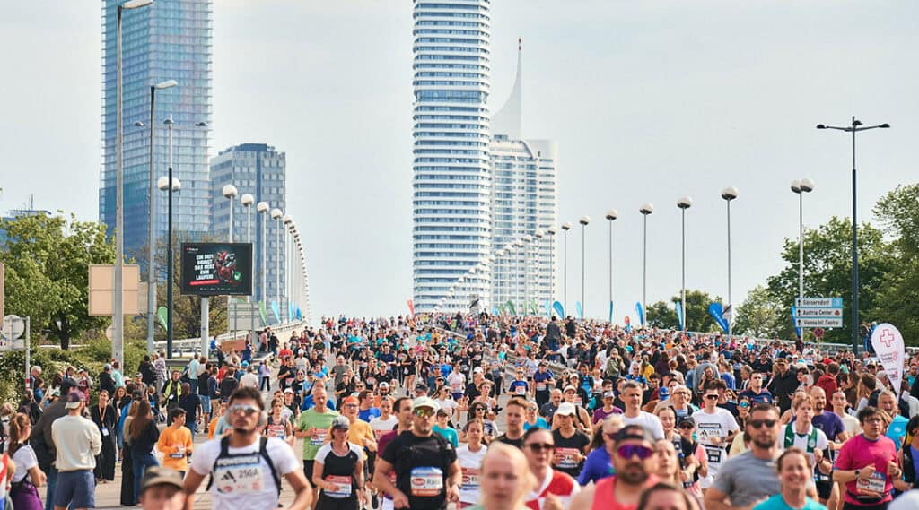 Großes Läuferfeld beim Vienna City Marathon auf einer breiten Straße, im Hintergrund die moderne Skyline von Wien.