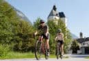 Zwei Radfahrer fahren auf einer Straße durch einen Ort in den Kitzbüheler Alpen, im Hintergrund eine Kirche und Berglandschaft.